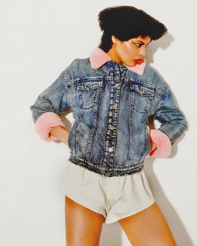 A woman poses in a denim jacket with faux fur trim and neutral shorts against a white background in a studio-style setup.