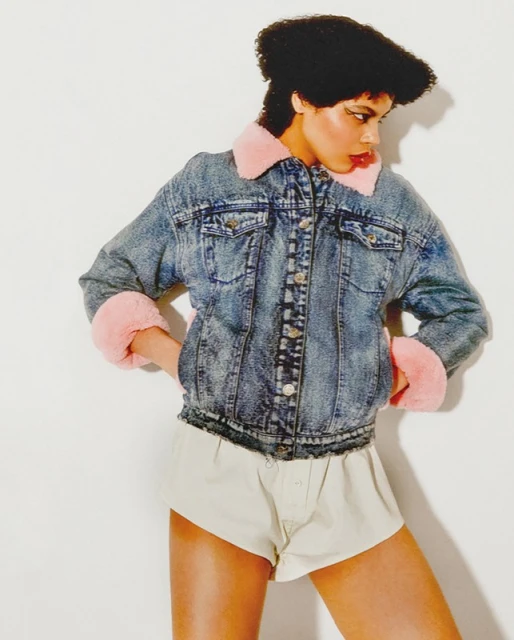 A woman poses in a denim jacket with faux fur trim and neutral shorts against a white background in a studio-style setup.