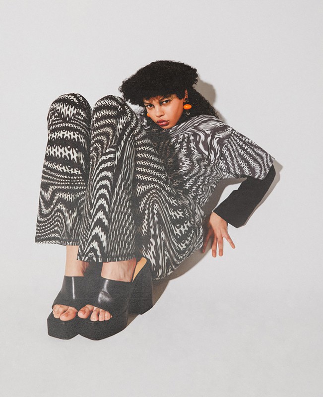 Fashion shot of a woman wearing a black-and-white patterned shirt and pants with platform shoes, posing in a photo studio with a white background.