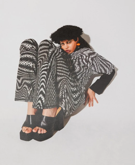 Fashion shot of a woman wearing a black-and-white patterned shirt and pants with platform shoes, posing in a photo studio with a white background.