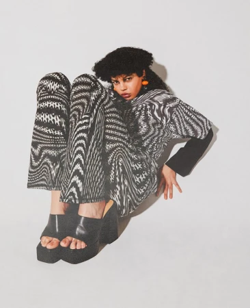 Fashion shot of a woman wearing a black-and-white patterned shirt and pants with platform shoes, posing in a photo studio with a white background.