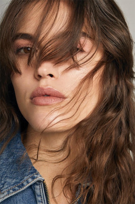 Close-up portrait of a young adult woman with brown hair partially covering her face, wearing a blue denim jacket against a neutral background.