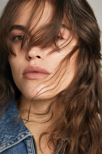 Close-up portrait of a young adult woman with brown hair partially covering her face, wearing a blue denim jacket against a neutral background.