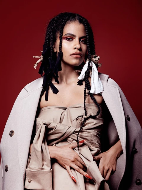 Portrait of a woman with long, braided hair posing in a beige coat and strapless dress against a deep red backdrop in a studio.