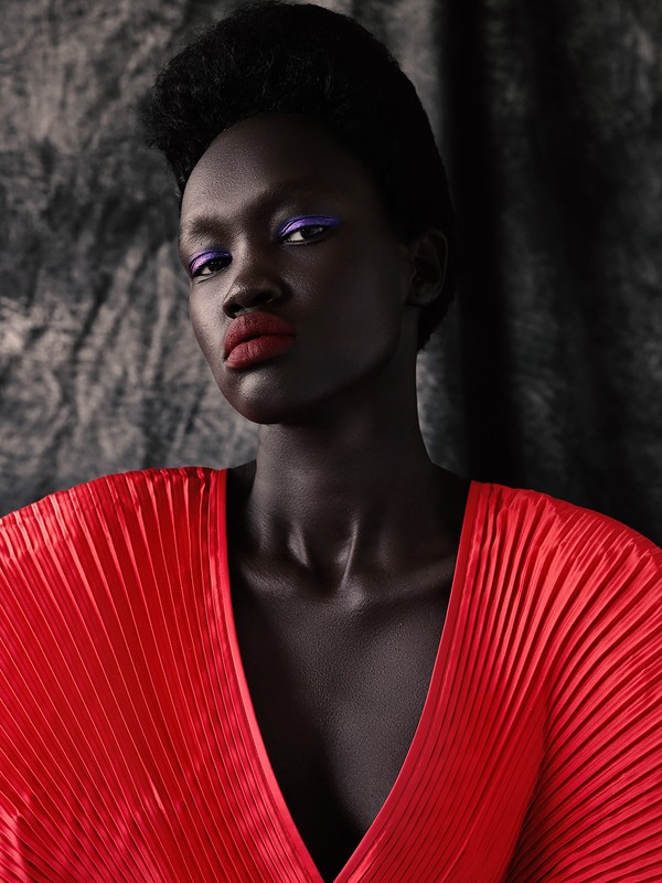 Elegant portrait of a woman with dark skin and bold makeup, wearing a red pleated top, set against a textured gray backdrop.