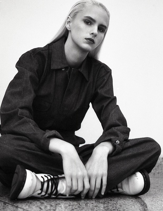 Young woman with platinum blonde hair poses in a denim jumpsuit and white boots against a white background, looking at the camera.