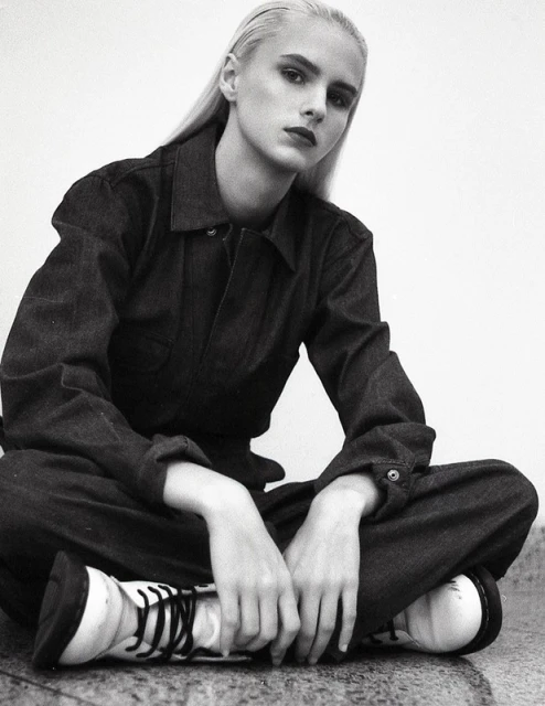 Young woman with platinum blonde hair poses in a denim jumpsuit and white boots against a white background, looking at the camera.
