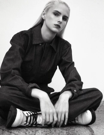 Young woman with platinum blonde hair poses in a denim jumpsuit and white boots against a white background, looking at the camera.