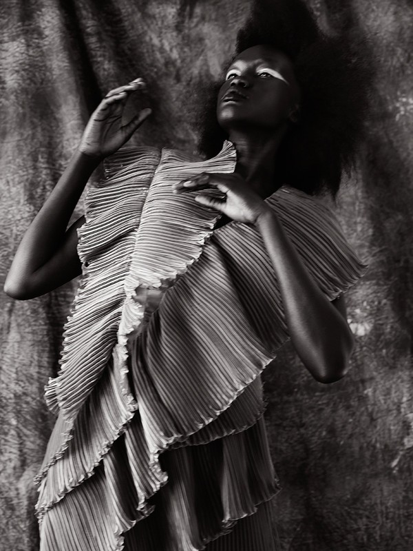 Monochrome portrait of a woman with dark hair and a pleated ruffle dress against a textured background, looking upwards.