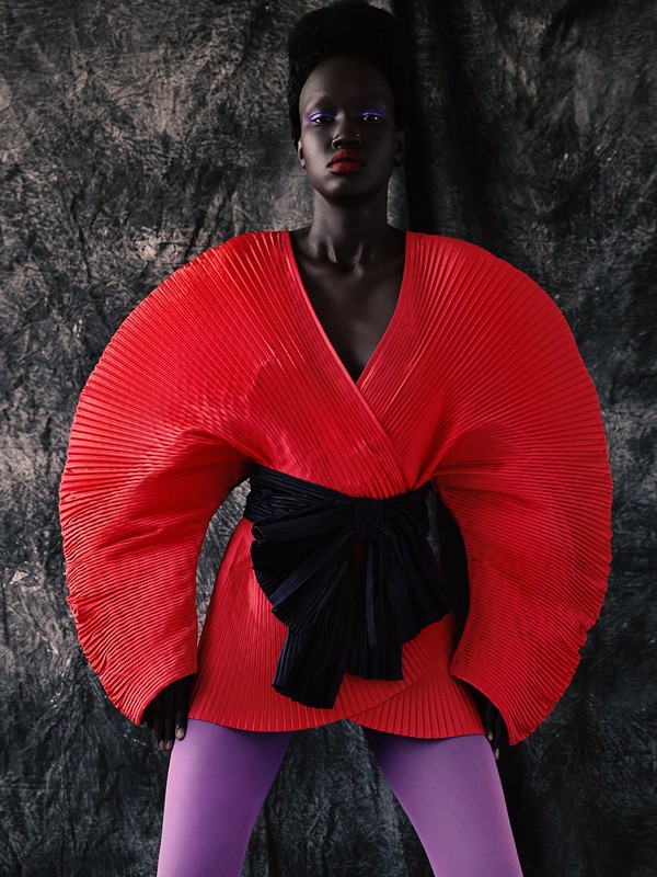 Woman in a red, pleated, wrap-style dress with a black obi belt and purple leggings, posing against a textured gray background.