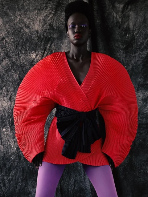 Woman in a red, pleated, wrap-style dress with a black obi belt and purple leggings, posing against a textured gray background.