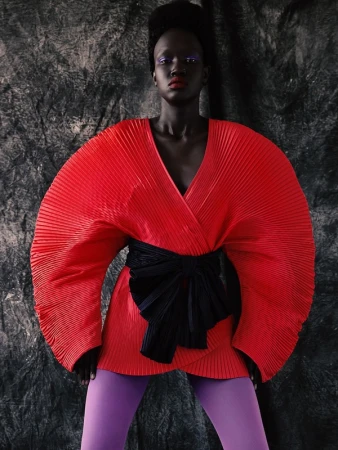 Woman in a red, pleated, wrap-style dress with a black obi belt and purple leggings, posing against a textured gray background.