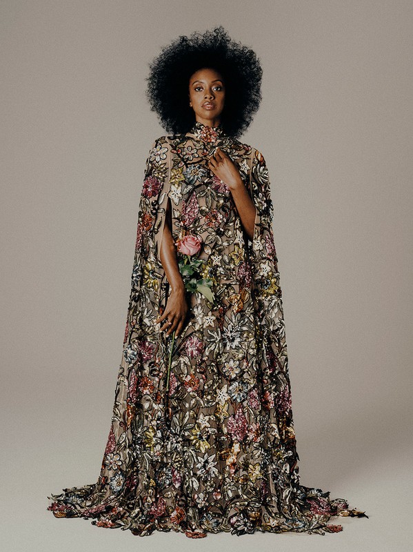 A woman with afro hairstyle in a floral embroidered cape gown holds a pink rose, posing against a plain grey background in a studio setting.