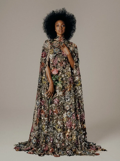 A woman with afro hairstyle in a floral embroidered cape gown holds a pink rose, posing against a plain grey background in a studio setting.