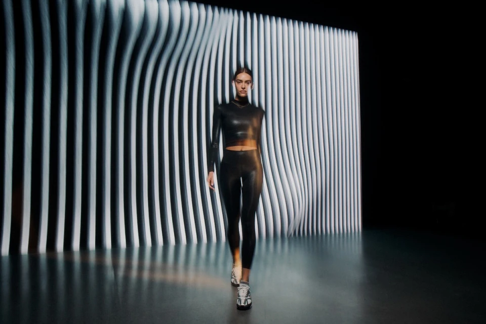 An adult female walking in black leggings and a black long-sleeved crop top against a black and white patterned background in a studio.