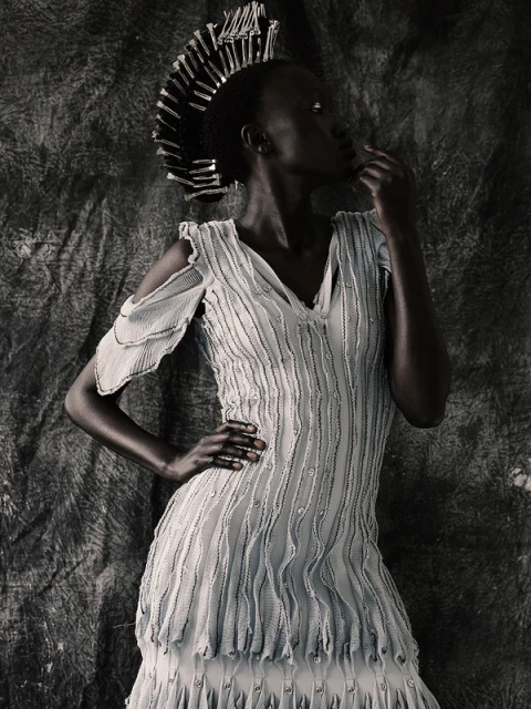 Portrait of a woman in a white dress and unique crown with hand on chin, against a dark textured backdrop. Fashion photography.