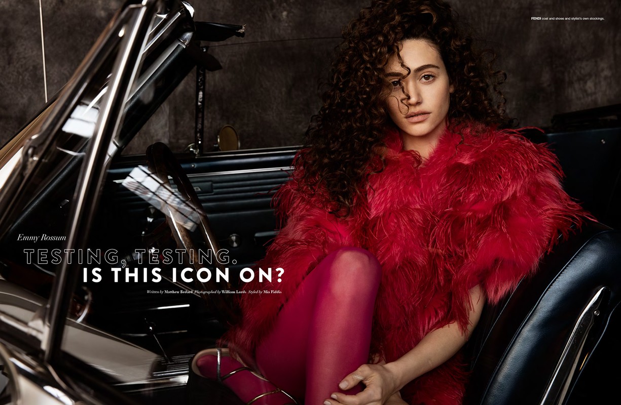 Emmy Rossum poses inside a classic car, wearing a striking red feathered coat and red stockings, creating a bold fashion statement in a studio setting.
