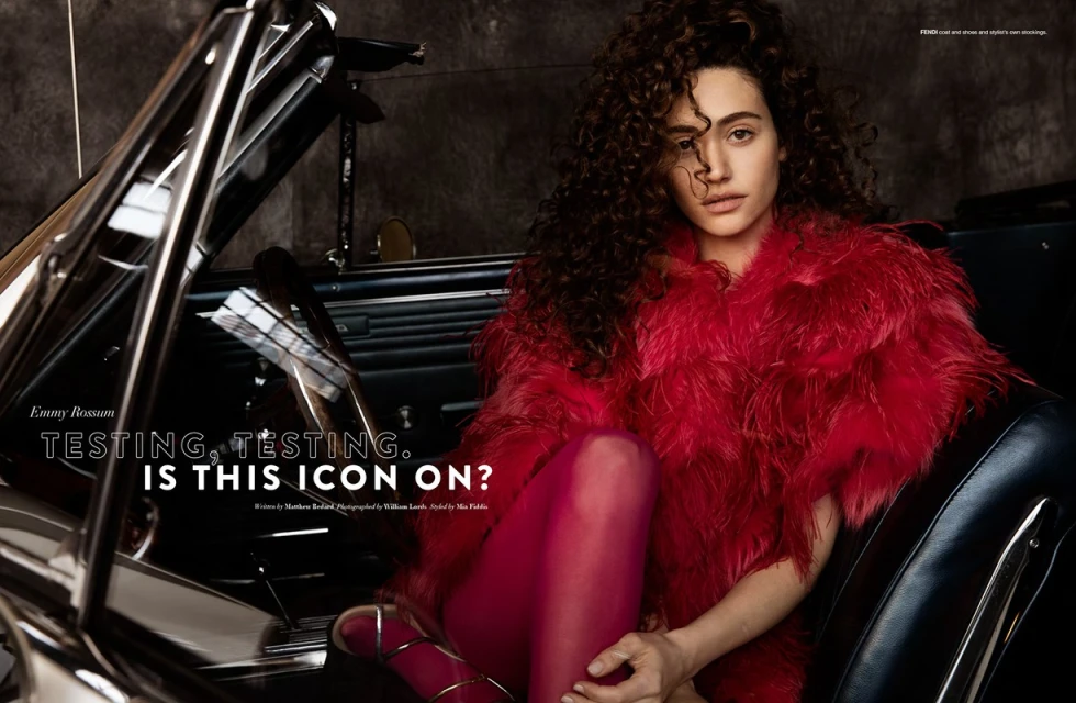 Emmy Rossum poses inside a classic car, wearing a striking red feathered coat and red stockings, creating a bold fashion statement in a studio setting.