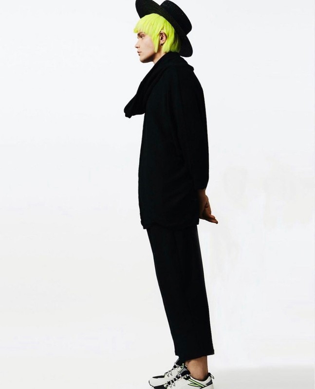 Side view of a person with neon green hair wearing black clothing, a hat, and sneakers against a white background in a studio setting.