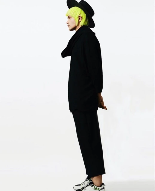 Side view of a person with neon green hair wearing black clothing, a hat, and sneakers against a white background in a studio setting.