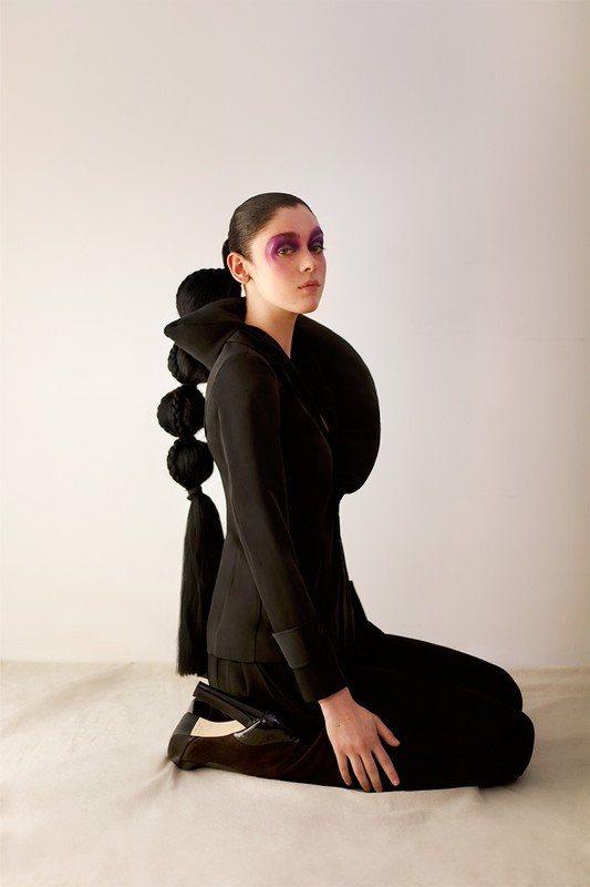 A young woman in a black suit kneels against a white backdrop, her face adorned with purple makeup and her hair styled in unique fashion.
