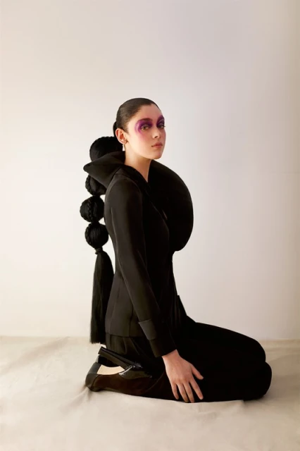 A young woman in a black suit kneels against a white backdrop, her face adorned with purple makeup and her hair styled in unique fashion.