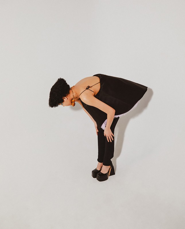 A woman bends at the waist with hands on knees, wearing a black top, pants, and chunky platform heels against a white background.