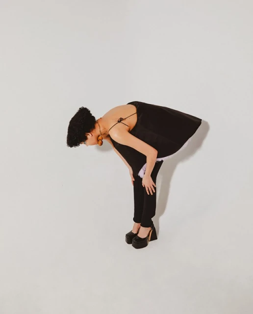 A woman bends at the waist with hands on knees, wearing a black top, pants, and chunky platform heels against a white background.