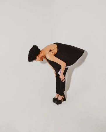 A woman bends at the waist with hands on knees, wearing a black top, pants, and chunky platform heels against a white background.