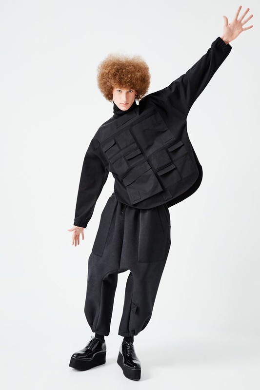Fashion model strikes a pose in a black oversized outfit, featuring a unique utility vest with pockets, wide-legged pants, and platform shoes.