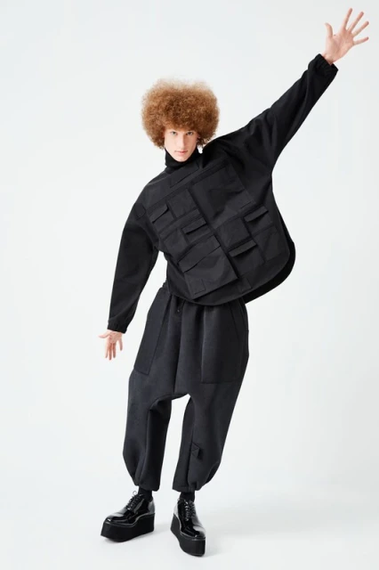 Fashion model strikes a pose in a black oversized outfit, featuring a unique utility vest with pockets, wide-legged pants, and platform shoes.