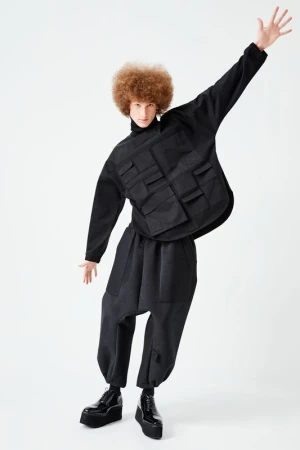 Fashion model strikes a pose in a black oversized outfit, featuring a unique utility vest with pockets, wide-legged pants, and platform shoes.