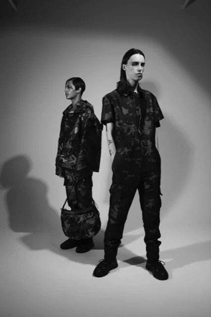 Two models in a black-and-white studio shot, both wearing dark camouflage-patterned outfits and shoes, with one carrying a matching bag.
