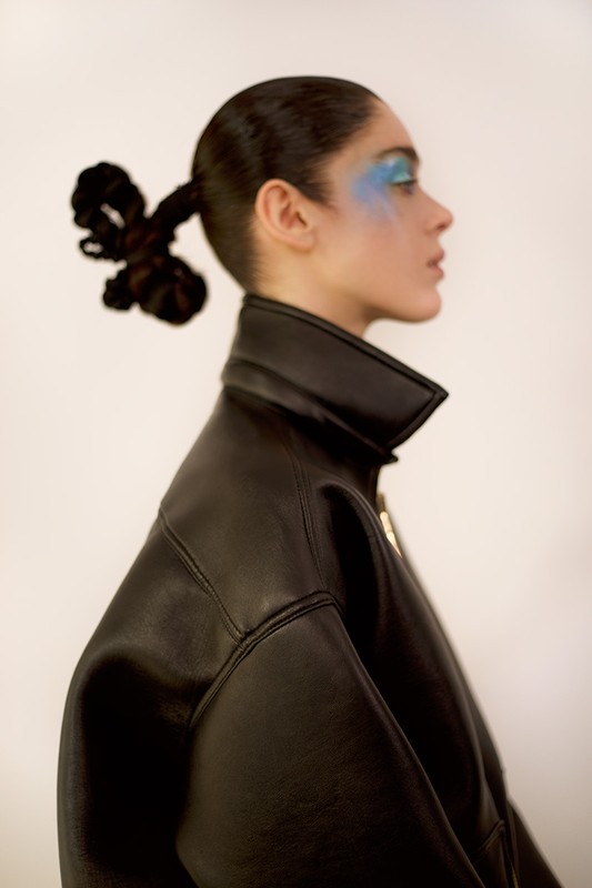 Profile of a woman with blue eyeshadow wearing a black leather jacket against a white background in a studio setting.