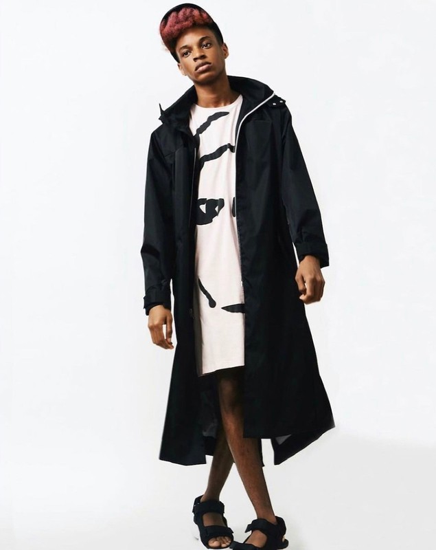 Young adult in black trench coat and sandals poses against a white backdrop for a fashion shoot with a confident expression.