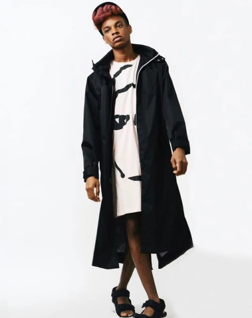 Young adult in black trench coat and sandals poses against a white backdrop for a fashion shoot with a confident expression.