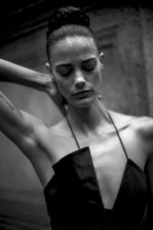 Monochrome portrait of a woman with her hair in a high bun, wearing a dark halter dress, posing with her hand behind her head.