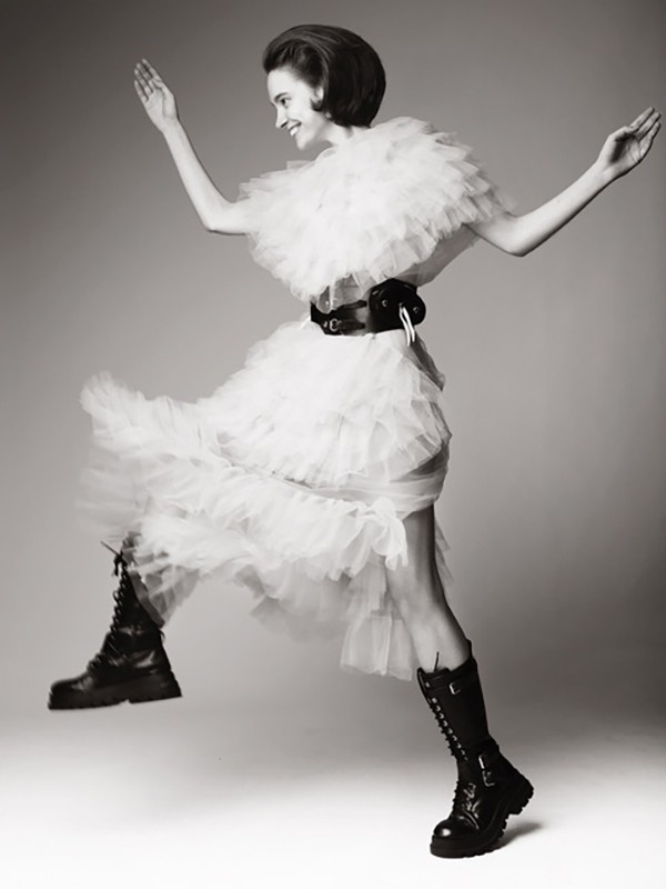 A fashionable woman in a white tulle dress and black combat boots strikes a joyful pose mid-air in a studio fashion shot.
