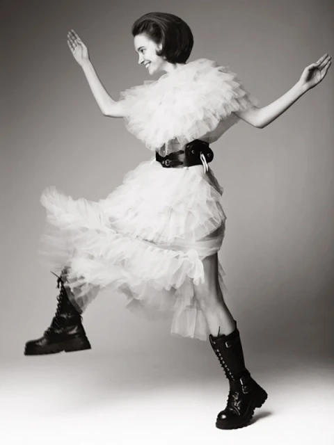 A fashionable woman in a white tulle dress and black combat boots strikes a joyful pose mid-air in a studio fashion shot.