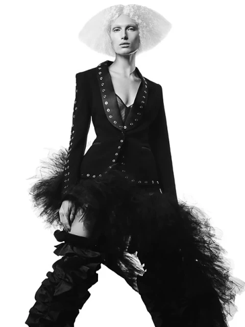 Monochrome portrait of a fashion model in a black jacket, tulle skirt, and statement white hair, posing against a white background.