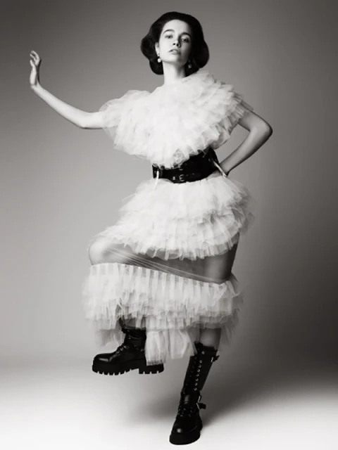 Fashion photo of a young woman posing in a tiered white tulle dress with a wide black belt and black boots, in a light grey studio.