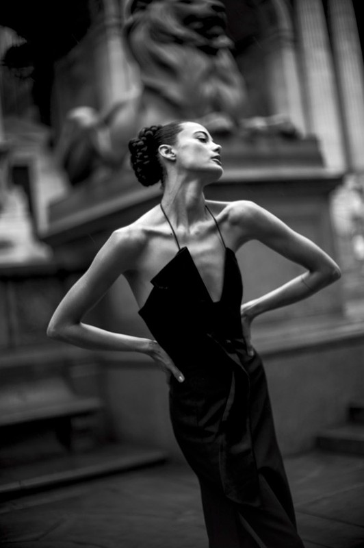 Monochrome fashion shot of a slender female model in a black dress posing in front of a stone lion sculpture in an outdoor setting.