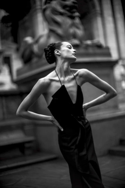 Monochrome fashion shot of a slender female model in a black dress posing in front of a stone lion sculpture in an outdoor setting.