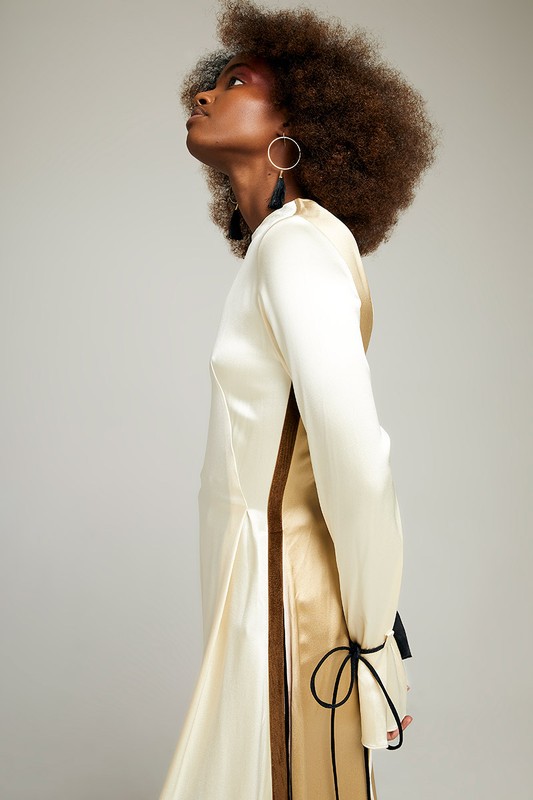 Elegant black woman with afro looking up, wearing a cream and gold dress with black details in a studio setting with grey background.