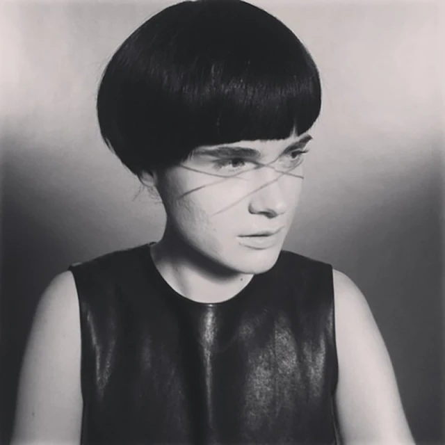 Portrait of a person with a bowl cut and sleeveless top in a photo studio setting. The shot is in black and white with grey background.