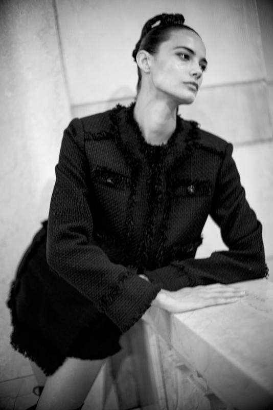 Black and white portrait of a woman in a black tweed suit, leaning on a white marble surface in an indoor setting.