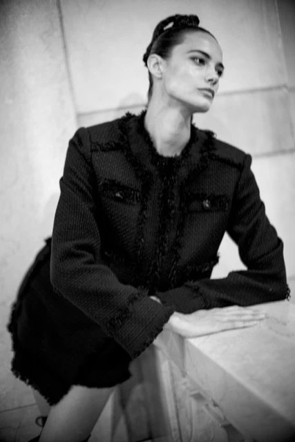 Black and white portrait of a woman in a black tweed suit, leaning on a white marble surface in an indoor setting.