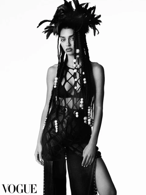 Monochrome portrait of a woman wearing a headdress with black feathers, beaded braids, a mesh top, and wide-legged slit pants.