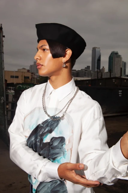 Young adult man poses in an urban environment, wearing a black hat, statement shirt, and chain necklace, with a cityscape background.
