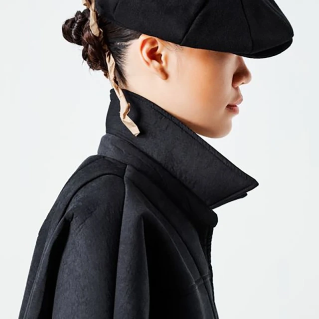 Side profile of an adult female wearing a black cap and coat against a white background in a studio setting with soft, diffused light.